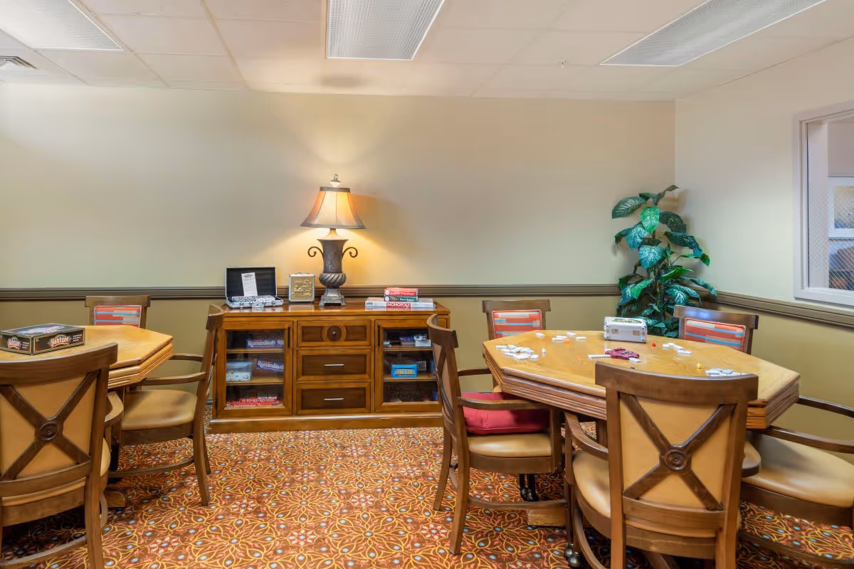 Interior activity room with wooden tables and chairs, a sideboard with a lamp and board games, and a potted plant.