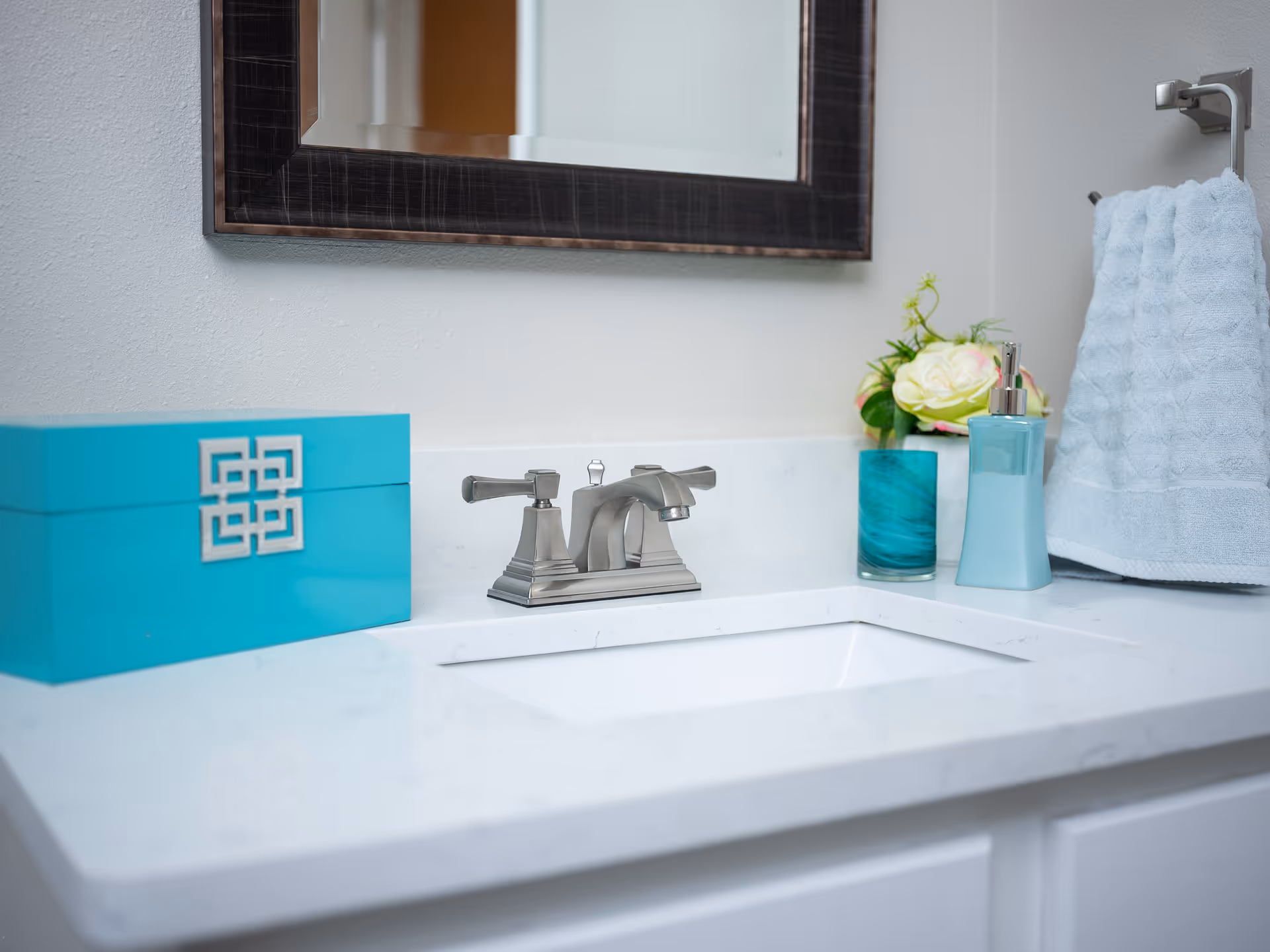 A bathroom sink countertop with a silver faucet, a turquoise decorative box, a blue soap dispenser, a blue glass holding flowers, and a light blue hand towel hanging on a silver towel ring. A large mirror with a dark wooden frame is mounted on the wall above the sink.