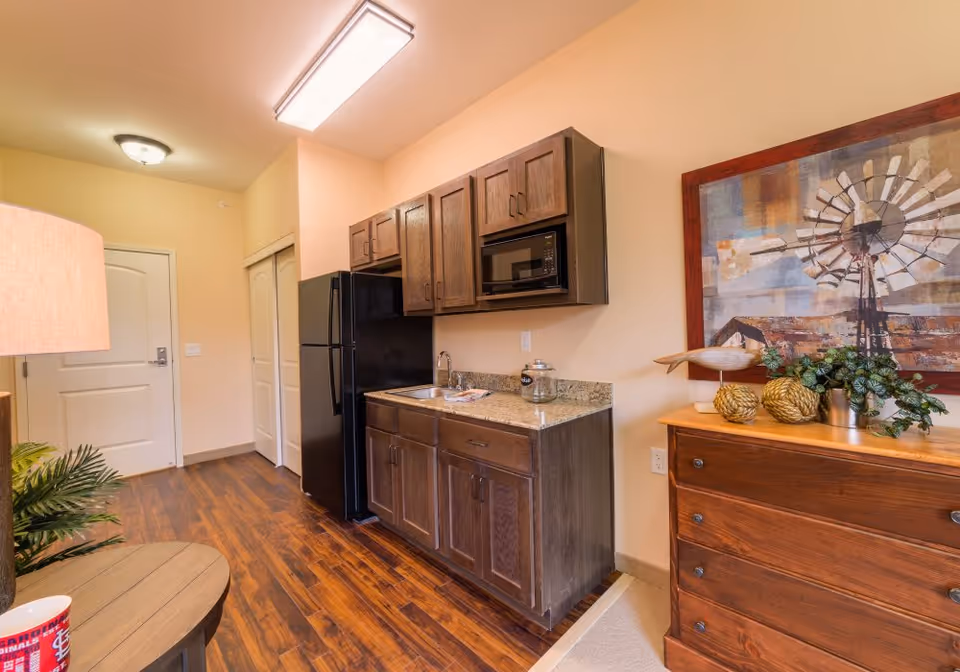 Small kitchenette and entry area with dark wood cabinets, black refrigerator and microwave, a sink and granite countertop, and a wooden dresser with decor and a framed windmill painting.