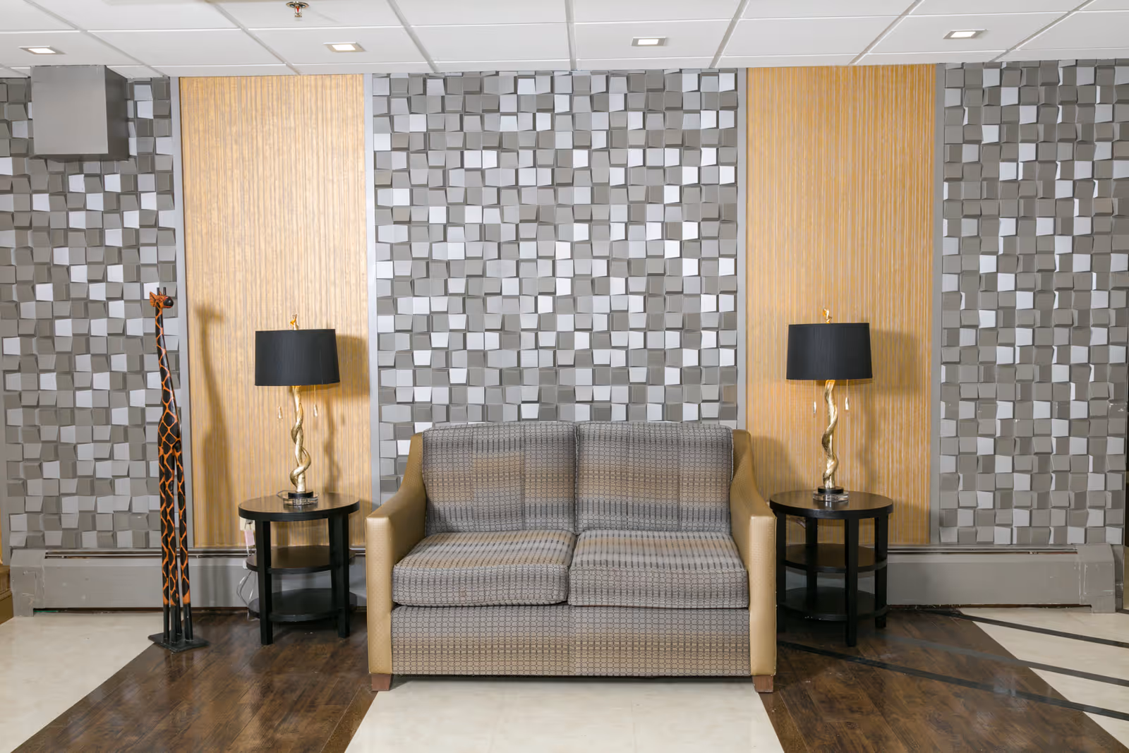 A modern seating area with a patterned two-seater sofa flanked by two black round side tables, each holding a lamp with a twisted gold base and black shade. The background features a textured wall with a geometric 3D pattern in gray and two vertical gold panels. A tall decorative wooden giraffe sculpture stands to the left of the sofa. The floor has a combination of dark wood and light tile.