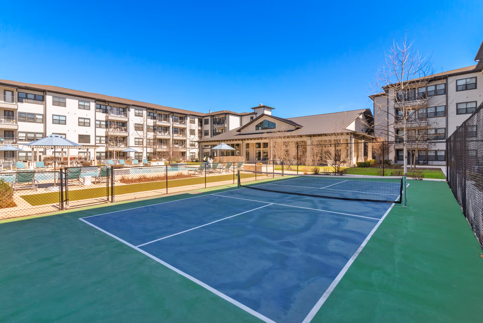 Outdoor view of a senior living courtyard with a fenced tennis/pickleball court in the foreground, a pool and umbrellas, and multi-story residential buildings in the background under a clear blue sky.