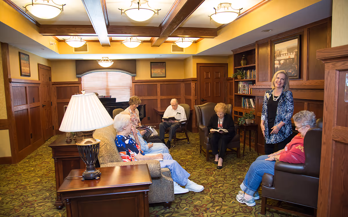 A group of elderly residents seated in a warmly lit communal living room, chatting and reading while a staff member stands smiling.