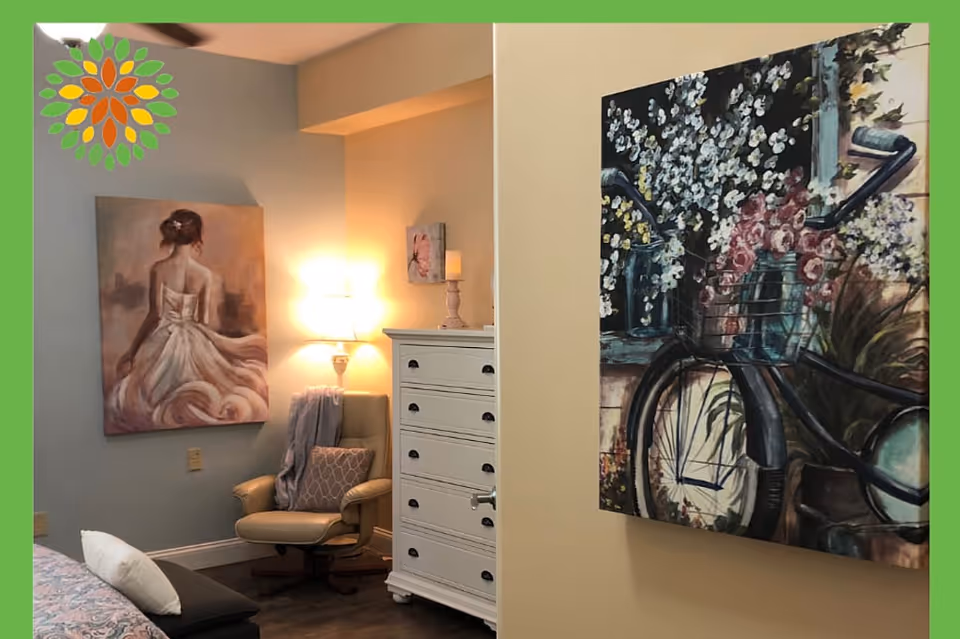 A cozy interior corner of a room featuring a beige armchair with a patterned pillow and a throw blanket, a white chest of drawers with a lamp and candle on top, and two paintings on the walls. One painting depicts a woman in a flowing dress seen from the back, and the other shows a bicycle basket filled with flowers.