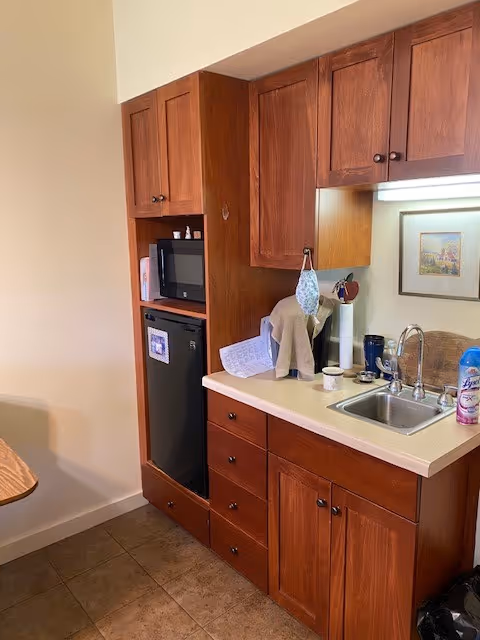 Small kitchenette with wooden cabinets, a sink, countertop, microwave, and mini-fridge.