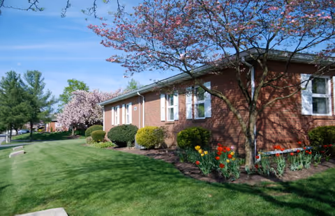 Exterior view of a single-story brick building with white-framed windows, surrounded by green grass, bushes, and blooming trees with pink flowers under a clear blue sky.