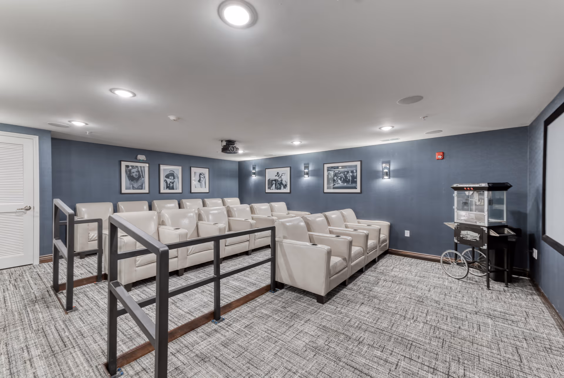 A small theater room with three rows of beige leather recliner chairs facing a large projection screen. The walls are painted blue and decorated with black and white framed photographs. There is a popcorn machine on the right side of the room and a door on the left side. The floor is carpeted in a gray pattern, and the ceiling has recessed lighting.