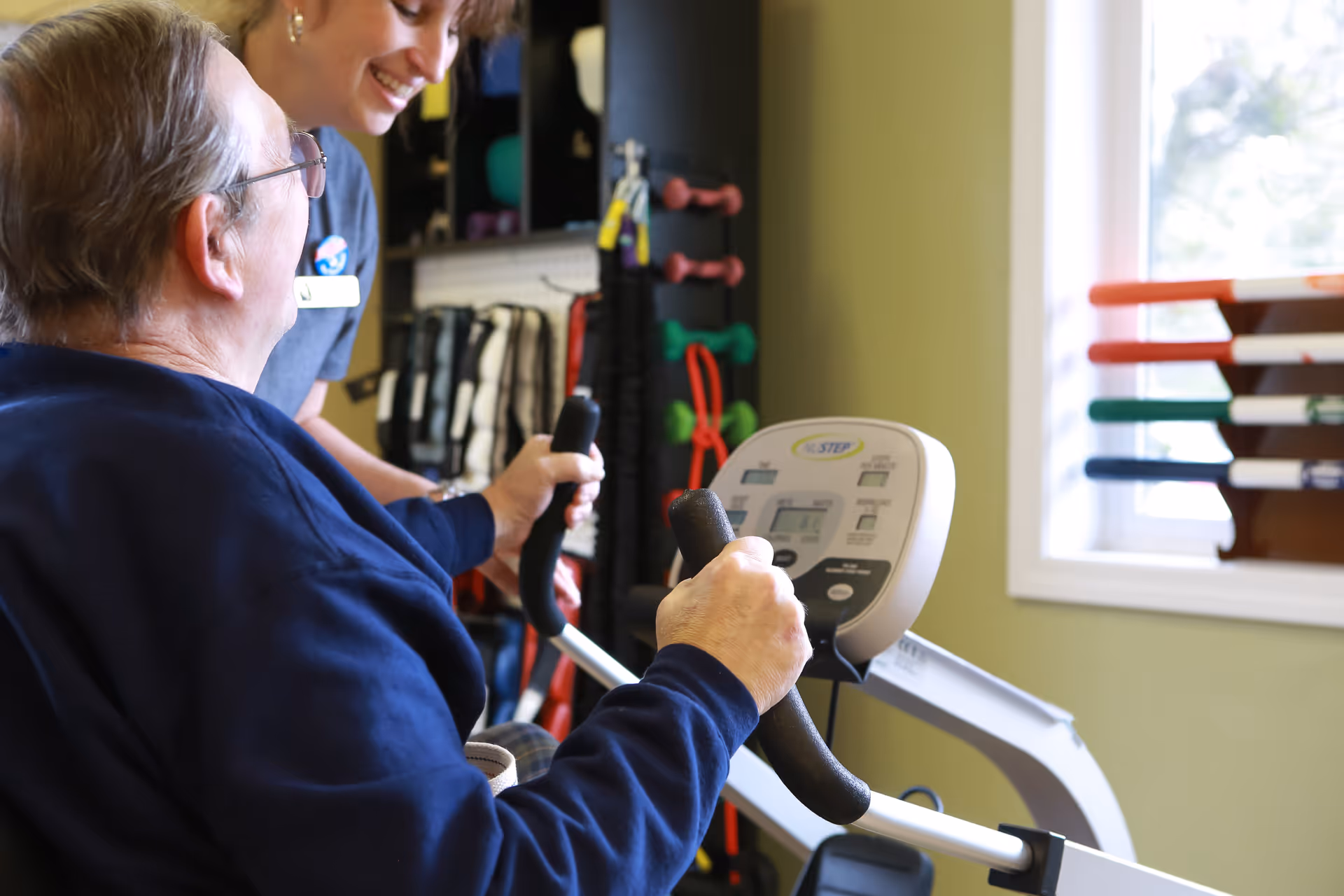 An elderly man using an exercise machine with the assistance of a smiling caregiver in a room with exercise equipment and a window.