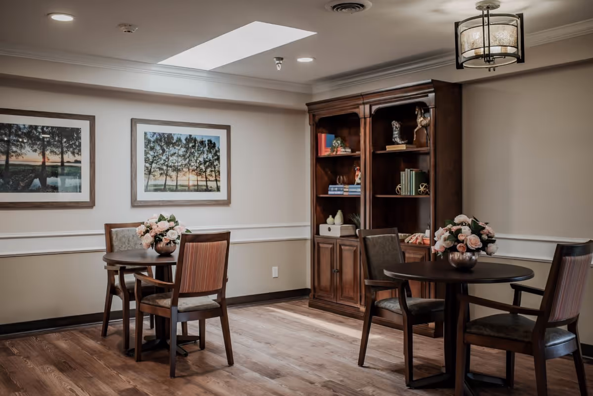 A cozy interior room with two round wooden tables, each surrounded by two chairs. Both tables have vases with pink and white flowers. On the wall, there are two framed landscape photographs of trees at sunset. A wooden bookshelf filled with books and decorative items stands against the wall. The room has wooden flooring, beige walls with white trim, and a ceiling light fixture.