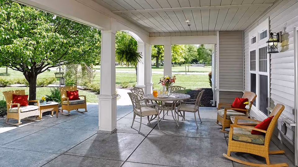 Covered outdoor patio area with wicker rocking chairs and a round metal table with chairs. The patio overlooks a green lawn with trees and shrubs. There are red cushions on the chairs and a vase with flowers on the table.
