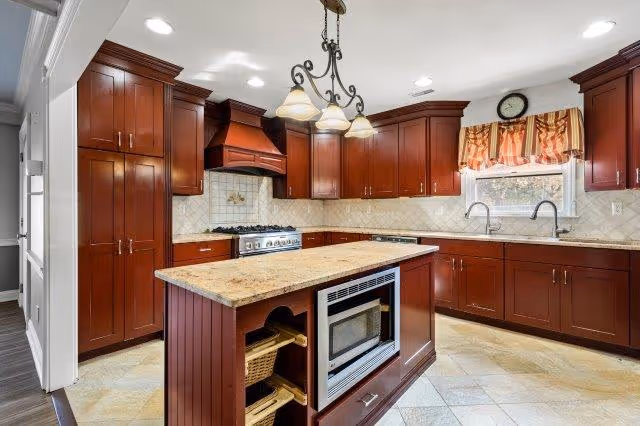 A spacious kitchen with dark wooden cabinets, a central island with a built-in microwave and storage baskets, beige tiled floor, and a window with a patterned valance above the sink. The kitchen features a stainless steel stove with a matching hood and a three-light chandelier hanging above the island.