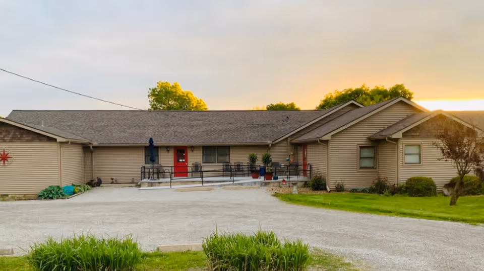 Exterior view of a single-story residential care center building with beige siding and a red front door, surrounded by greenery and a gravel driveway under a cloudy sky at sunset.