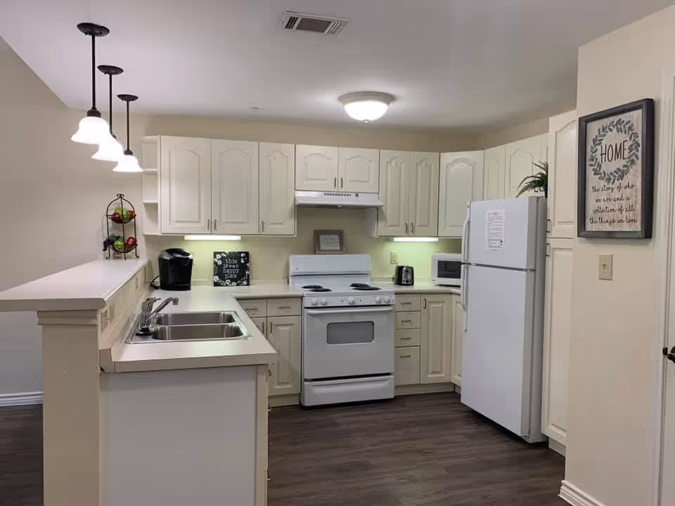 A clean and well-lit kitchen with white cabinets and appliances including a stove, refrigerator, microwave, and toaster. There is a double sink on the counter with a coffee maker and a three-tier fruit basket on the left side. The kitchen has dark wood flooring and pendant lights hanging above the counter. A framed decorative sign is on the wall to the right.