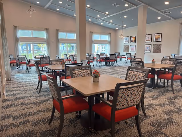 Dining room with multiple square tables and patterned chairs with red seats, large windows, and a decorative ceiling.