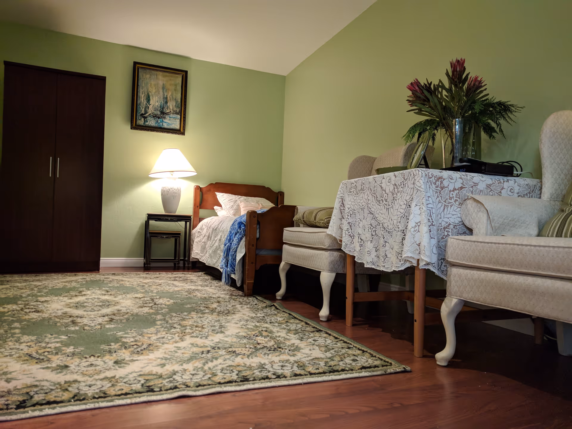 A cozy bedroom with light green walls, a wooden bed with white bedding and a blue throw blanket, a bedside table with a lamp, a dark wooden wardrobe, a patterned area rug, and a small table covered with a lace tablecloth holding a vase of flowers and some items. Two upholstered armchairs are positioned near the table.