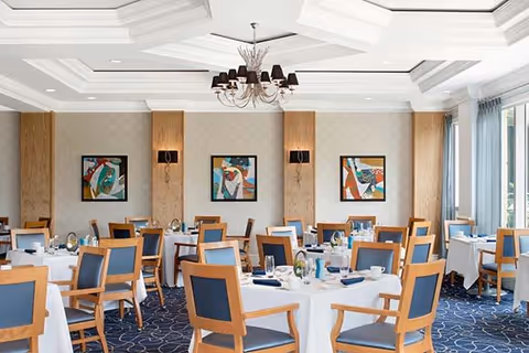 A bright dining room with round tables covered in white tablecloths, set with plates, glasses, and napkins. Wooden chairs with blue cushions surround the tables. The room features a patterned blue carpet, three abstract paintings on the wall, and a chandelier hanging from a coffered ceiling. Large windows with light curtains allow natural light to fill the space.