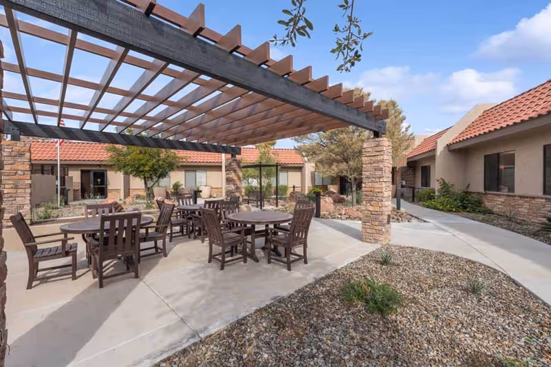 Courtyard with a wooden pergola shading round tables and chairs between single-story buildings with red tile roofs.