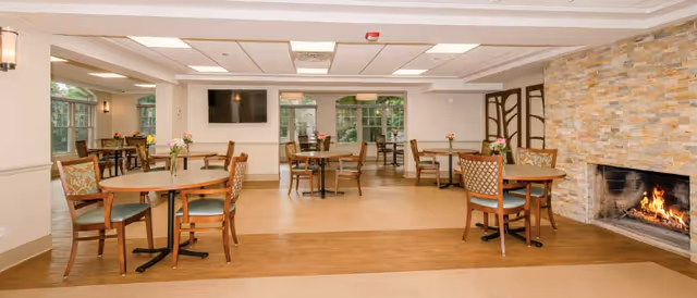 A spacious dining room with several round wooden tables and chairs arranged neatly. The room features large windows allowing natural light to enter, a stone fireplace with a fire burning, and a flat-screen TV mounted on the wall. The decor includes light-colored walls, ceiling lights, and small flower arrangements on the tables.