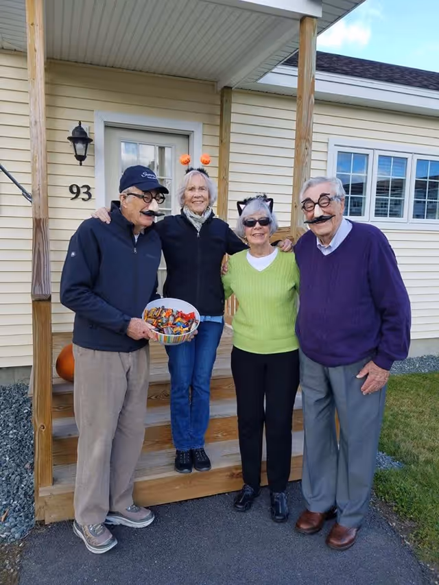 Four elderly people standing together outside a house with beige siding. They are wearing Halloween-themed accessories such as fake mustaches, glasses, cat ears, and pumpkin headbands. One person is holding a bowl filled with candy. The house has a white door with the number 93 next to it and a small wooden porch with steps.