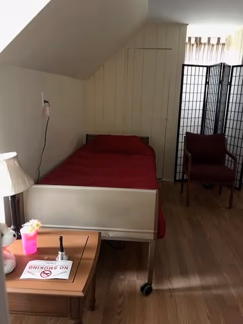 A small bedroom with a single bed covered in a red blanket and pillow. Next to the bed is a wooden nightstand with a lamp, a pink cup, a small teddy bear, and a 'No Smoking' sign. There is a brown chair and a black folding screen near the window with sheer curtains. The room has wooden flooring and slanted white walls.