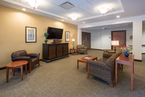 A cozy living room area in a senior living facility with beige walls and carpeted floor. The room features a flat-screen TV mounted on a wooden cabinet, two framed pictures on the wall, a small round wooden table with two leather chairs, a patterned sofa, a wooden coffee table with magazines, a side table with a lamp and a blue vase with flowers, and an armchair in the background near a door.