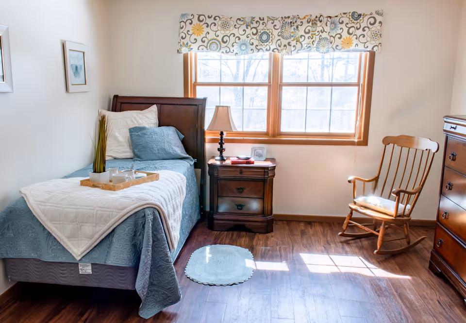 Sunlit bedroom with a single bed, nightstand and lamp by a window, a wooden rocking chair, and hardwood floors.