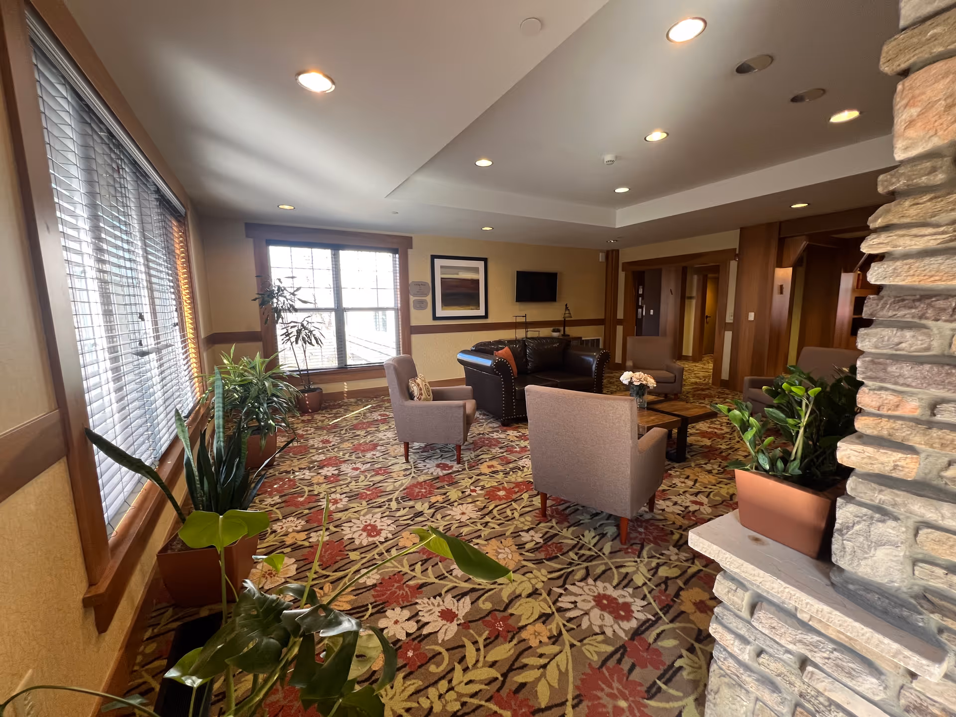 A cozy living room area in Deer Creek Lodge with a floral patterned carpet, several potted plants, a large window with blinds, a dark leather sofa, three beige armchairs around a wooden coffee table with a flower vase, a wall-mounted TV, and a stone fireplace on the right side.