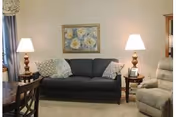 Cozy living room with a dark sofa flanked by two table lamps, a recliner, side tables, and a floral painting on the wall.