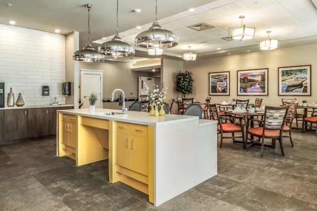 A spacious dining area in a senior living community featuring a large kitchen island with a sink and yellow cabinetry. The room has multiple dining tables with chairs that have orange cushions. The walls are decorated with framed pictures of boats on water, and there are modern pendant lights hanging above the kitchen island. The floor is covered with a dark tile, and there is a plant in the corner near the dining tables.