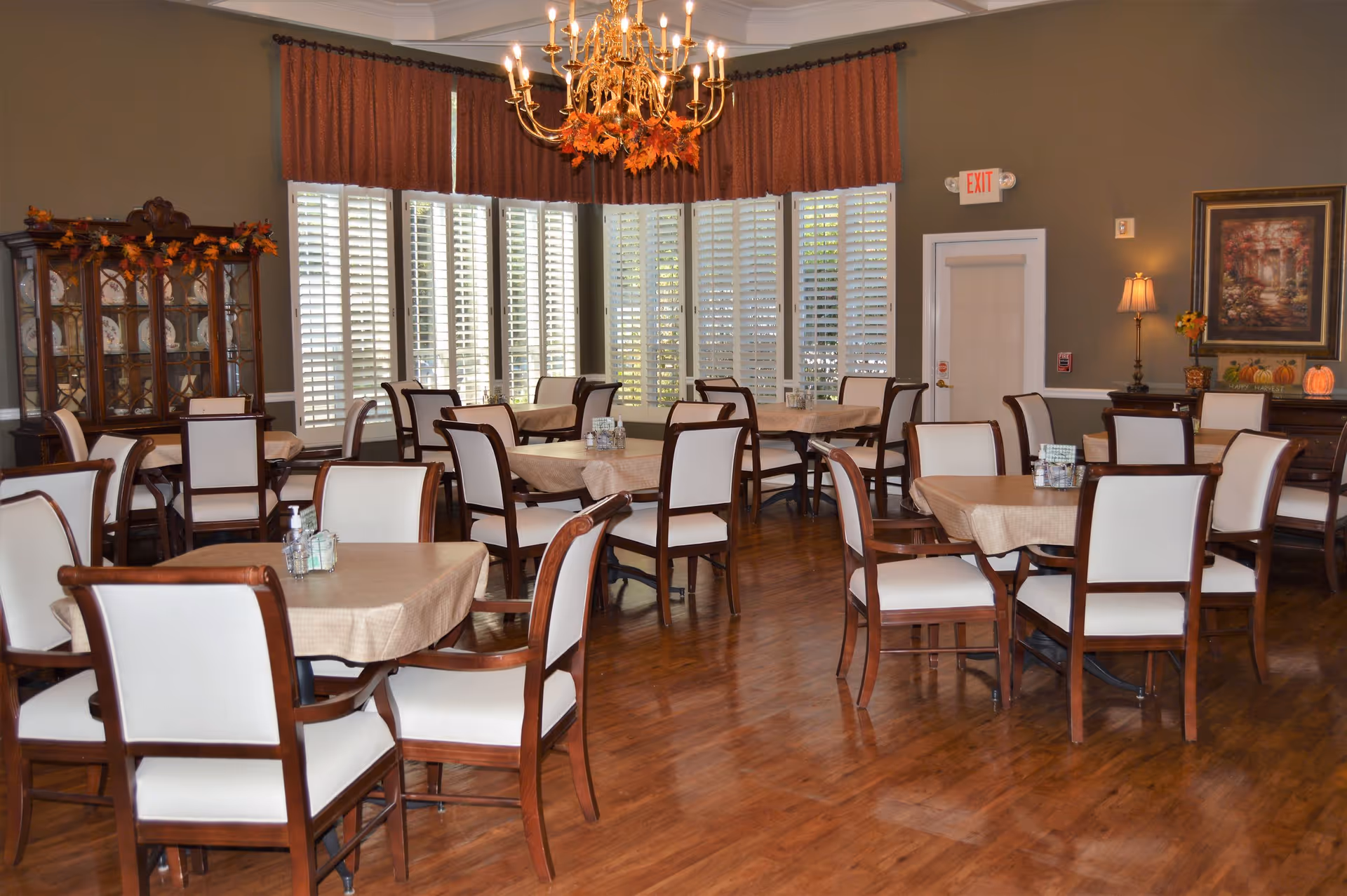 Formal dining room with multiple tables and upholstered chairs, a chandelier, and large shuttered windows.