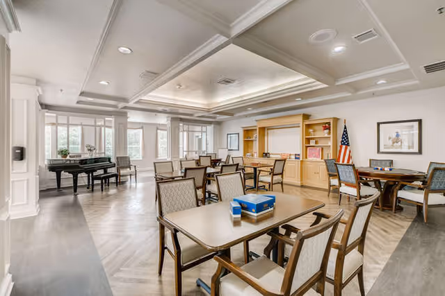A spacious, well-lit common area in a senior living facility with multiple tables and chairs arranged for group activities or dining. There is a grand piano near the windows, a built-in wooden shelving unit with decorations and an American flag, and framed artwork on the walls. The room has a light color scheme with wood flooring and recessed lighting in the ceiling.