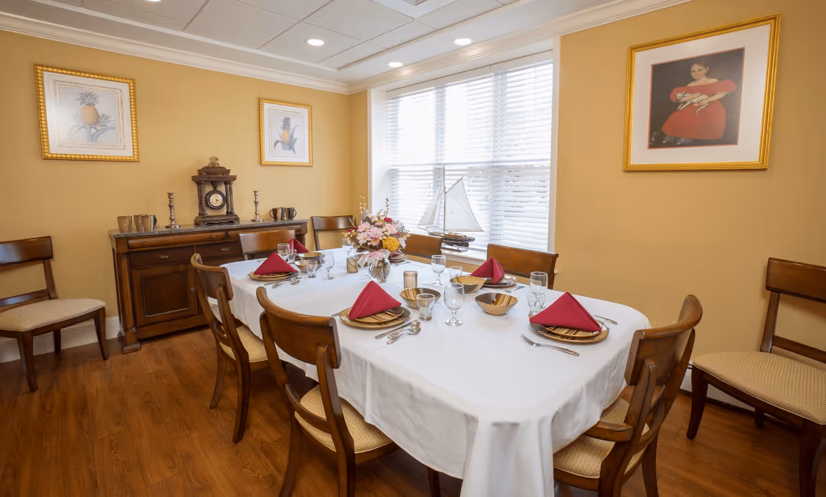 A dining room with a wooden table covered by a white tablecloth set for six people with red napkins, gold plates, and glassware. The room has wooden chairs, a wooden sideboard with a clock and decorative items, three framed pictures on the walls, and a window with blinds letting in natural light.