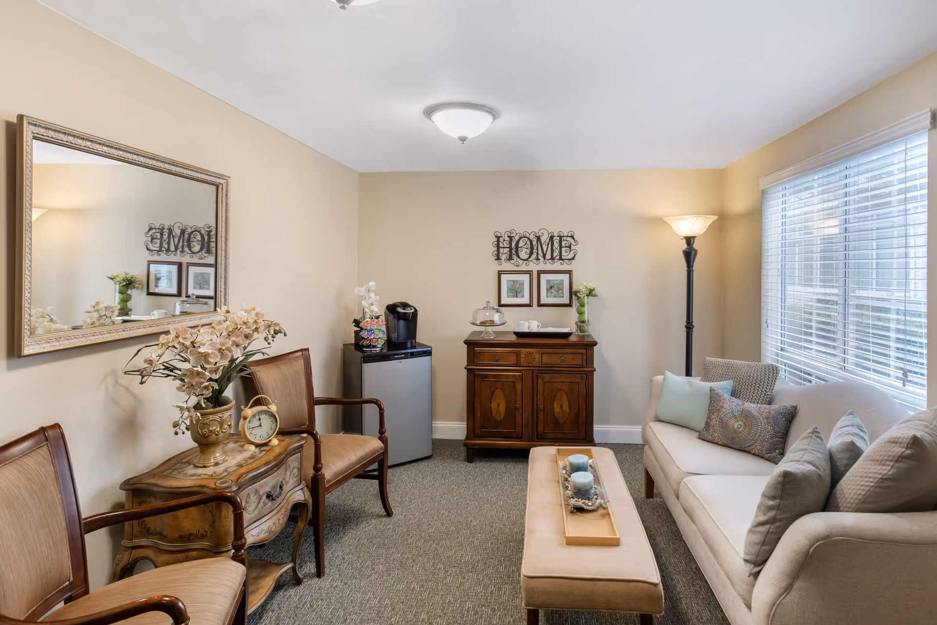A cozy living room with a beige sofa adorned with multiple decorative pillows, a cushioned bench with candles on a tray, two wooden armchairs with beige upholstery, a small ornate wooden table with a flower vase and a clock, a wooden cabinet with cups and a cake stand, a mini fridge with a coffee maker on top, a floor lamp, and a large window with blinds letting in natural light. A decorative wall piece spelling 'HOME' and two framed pictures hang above the cabinet.