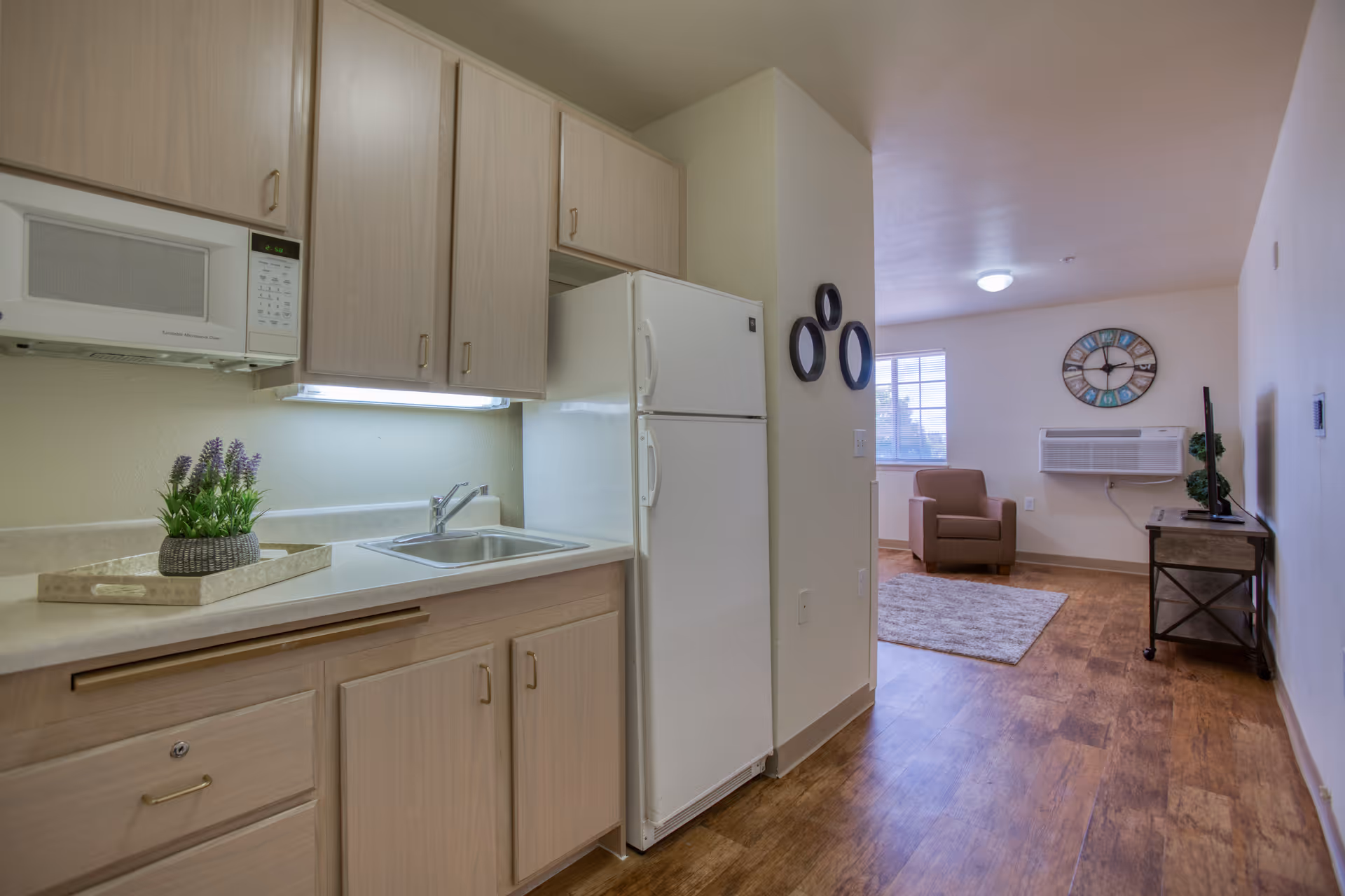 Interior view of a senior living facility apartment showing a small kitchen area with light wood cabinets, a white microwave, refrigerator, and a sink. A decorative tray with a small plant is on the countertop. Beyond the kitchen is a living area with a brown armchair, a wall-mounted air conditioning unit, a large decorative clock on the wall, and a TV on a stand. The floor is wood-style and the walls are light-colored.