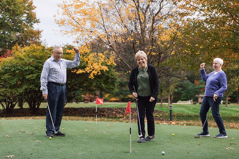 Three elderly people playing golf on a putting green outdoors surrounded by trees with autumn foliage. Two women and one man are smiling and appear to be celebrating a successful putt.