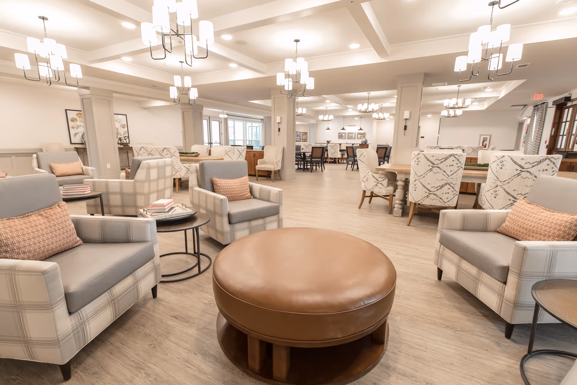 Spacious and well-lit common area with multiple armchairs featuring plaid upholstery and orange accent pillows, round leather ottoman in the center, several tables and chairs in the background, modern chandeliers hanging from the ceiling, and light wood flooring.
