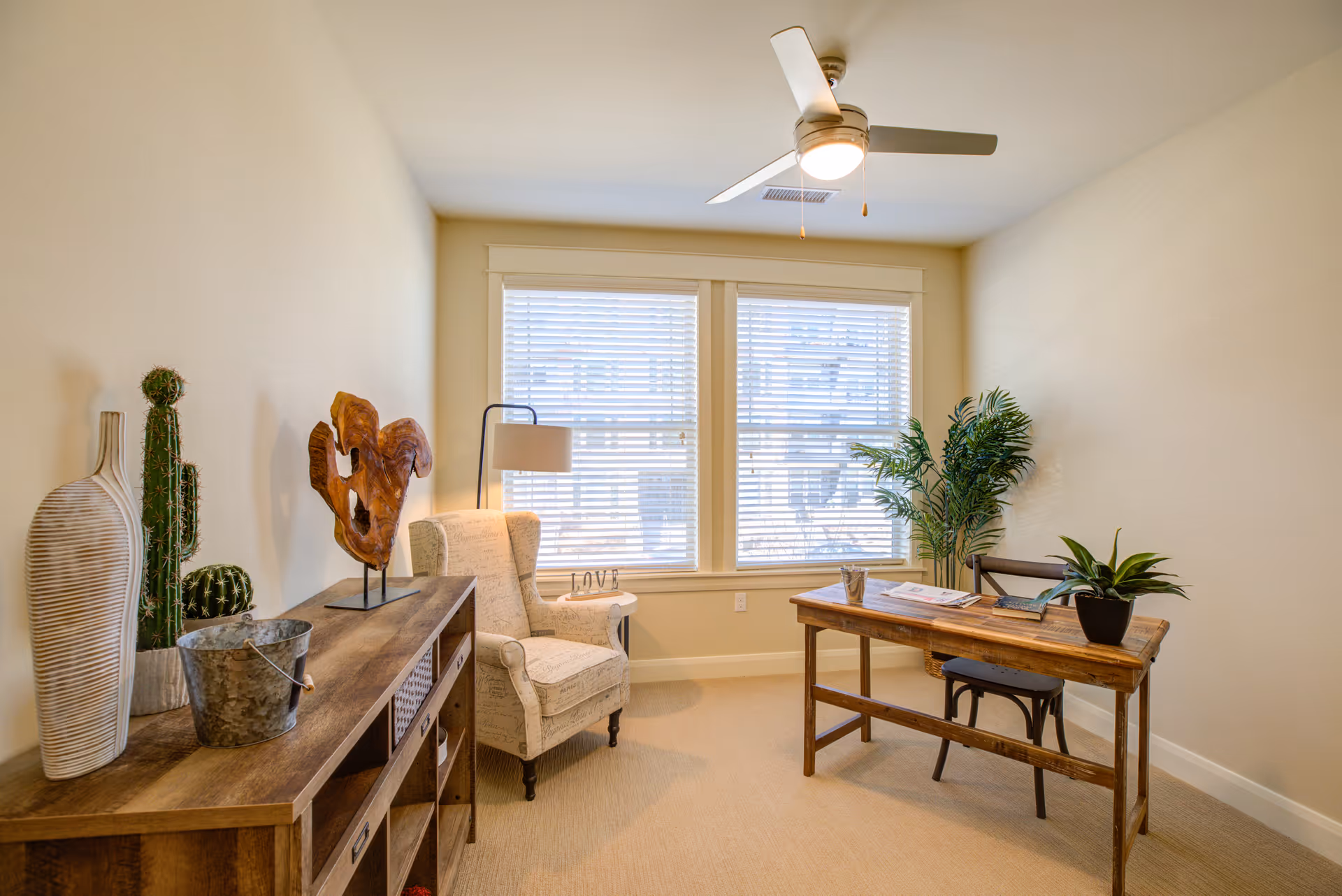 A bright room with beige walls and carpet featuring a wooden desk with a small potted plant, books, and a metal container on top. There is a wooden chair behind the desk and a large potted plant in the corner near two windows with white blinds. On the left side, there is a wooden console table with decorative items including a tall white vase, a metal bucket, a cactus plant, and a wooden sculpture. An upholstered armchair with script pattern and a floor lamp are positioned near the windows. A ceiling fan with a light is mounted on the ceiling.