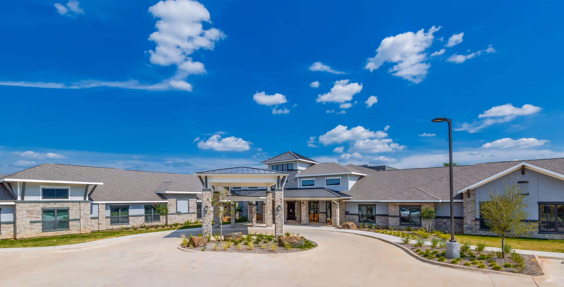 Front exterior view of Lynridge of Arlington Assisted Living & Memory Care facility under a bright blue sky with scattered clouds. The building features a covered entrance with stone pillars, multiple windows, and a well-maintained driveway with landscaped greenery.