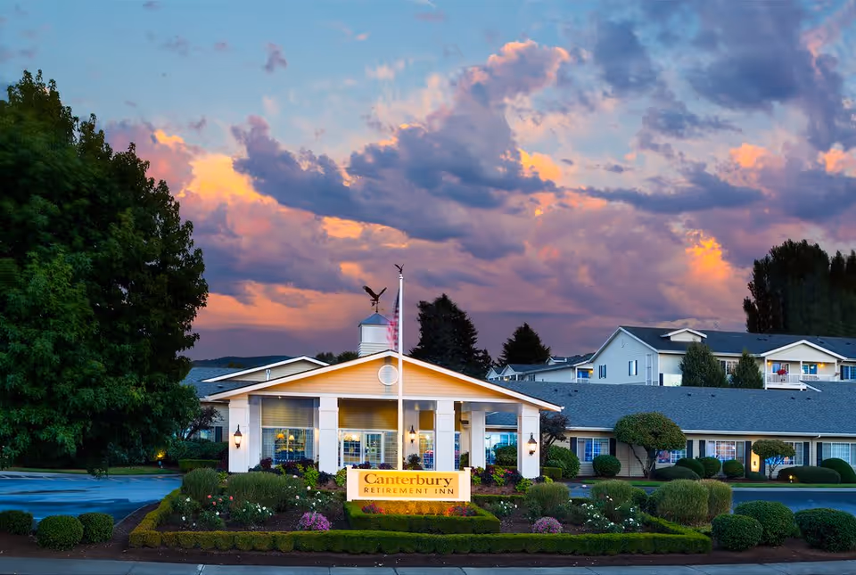 Front entrance of the Canterbury Inn assisted living community with landscaped gardens, a lit sign, and a dramatic sunset sky.