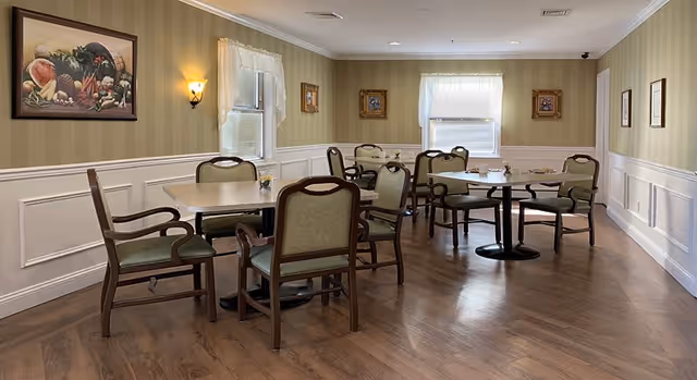 A dining room with several tables and chairs arranged neatly. The room has wood flooring, striped wallpaper with a chair rail, framed pictures on the walls, and two windows with sheer white curtains letting in natural light.