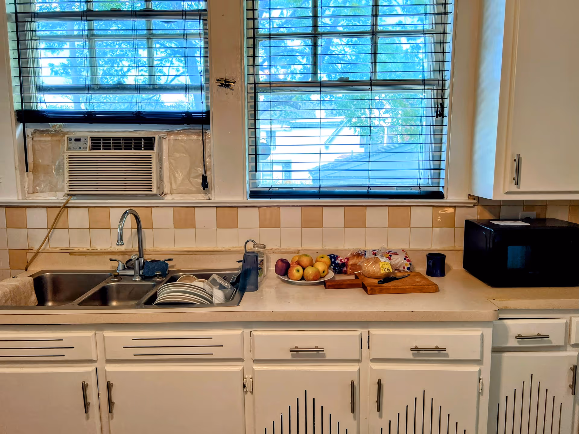 A kitchen countertop with a double sink filled with clean dishes, a plate of apples, a loaf of bread on a cutting board with a knife, a blue mug, and a black microwave. Two windows with blinds are above the counter, one with a window air conditioning unit installed.