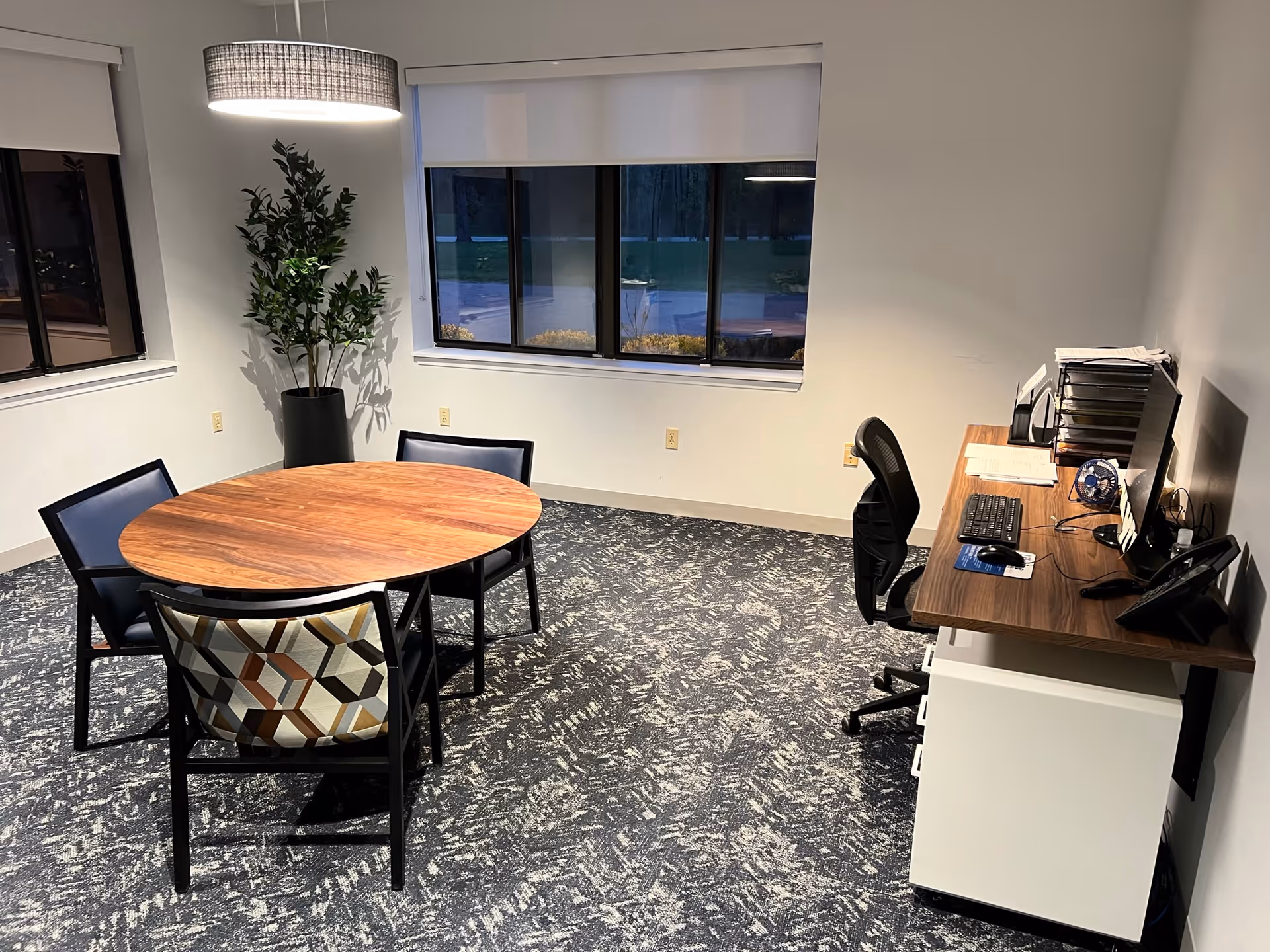 Interior meeting/office room with a round wooden table and chairs, a desk with a computer, and a potted plant under a pendant light.