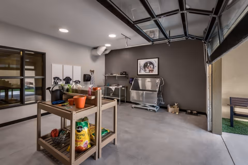 A clean, well-lit indoor space with a gardening workbench holding pots, gloves, and potting mix. The room has a large open garage-style door leading outside to a small bench and artificial grass. On the far wall, there is a stainless steel pet washing station, a shelf with grooming supplies, and a framed picture of dogs. The floor is concrete and the walls are painted in neutral colors.