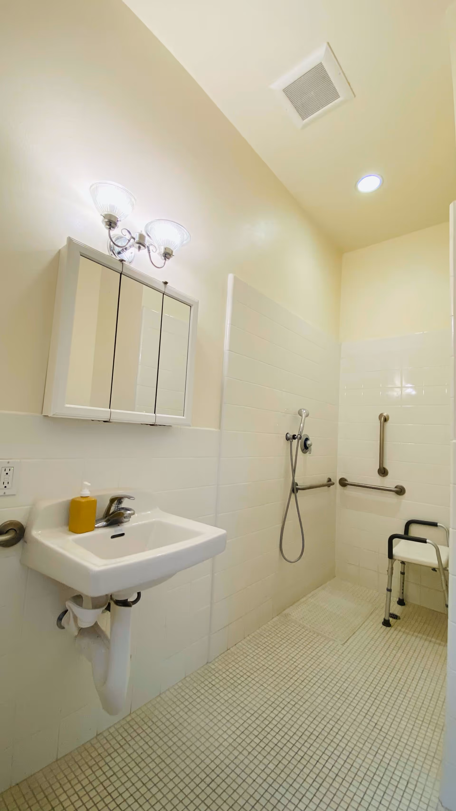 A clean, accessible bathroom with a wall-mounted sink, a mirror cabinet above it, a soap dispenser, and a walk-in shower area equipped with a handheld showerhead and grab bars. There is also a shower chair present. The walls and floor are tiled in light colors, and the room is well-lit with ceiling and wall lights.