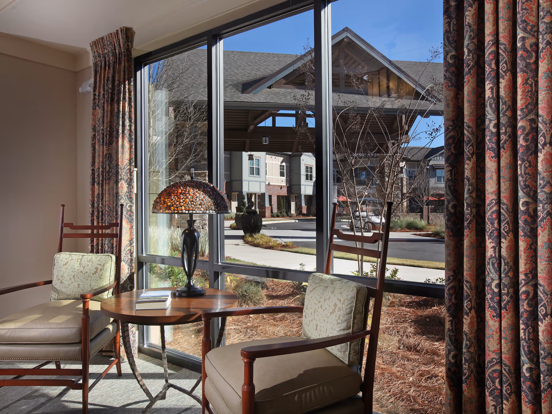 A cozy seating area with two cushioned wooden chairs and a round wooden table with a decorative lamp on it, positioned next to large windows with patterned curtains. Outside the window, a covered entrance and part of the building exterior are visible under a clear blue sky.
