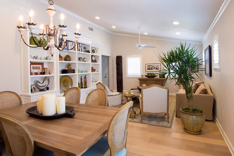 Open-plan dining and living room with a wooden dining table and chandelier, built-in shelving, seating area and a large potted plant.