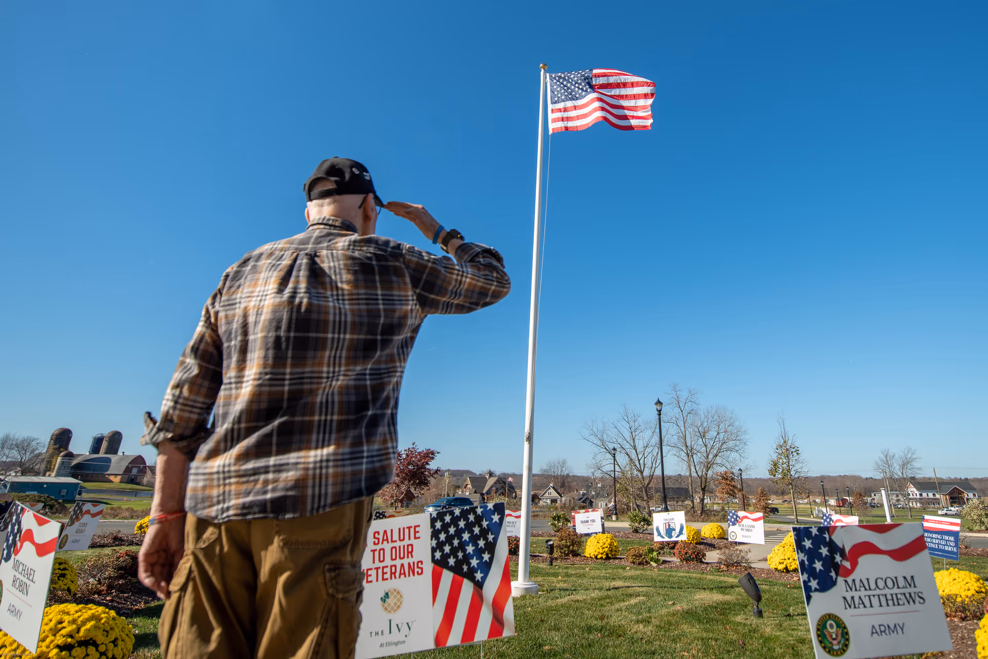 An elderly man wearing a plaid shirt and cap salutes an American flag flying on a flagpole in a memorial garden with signs honoring veterans and yellow flowers.