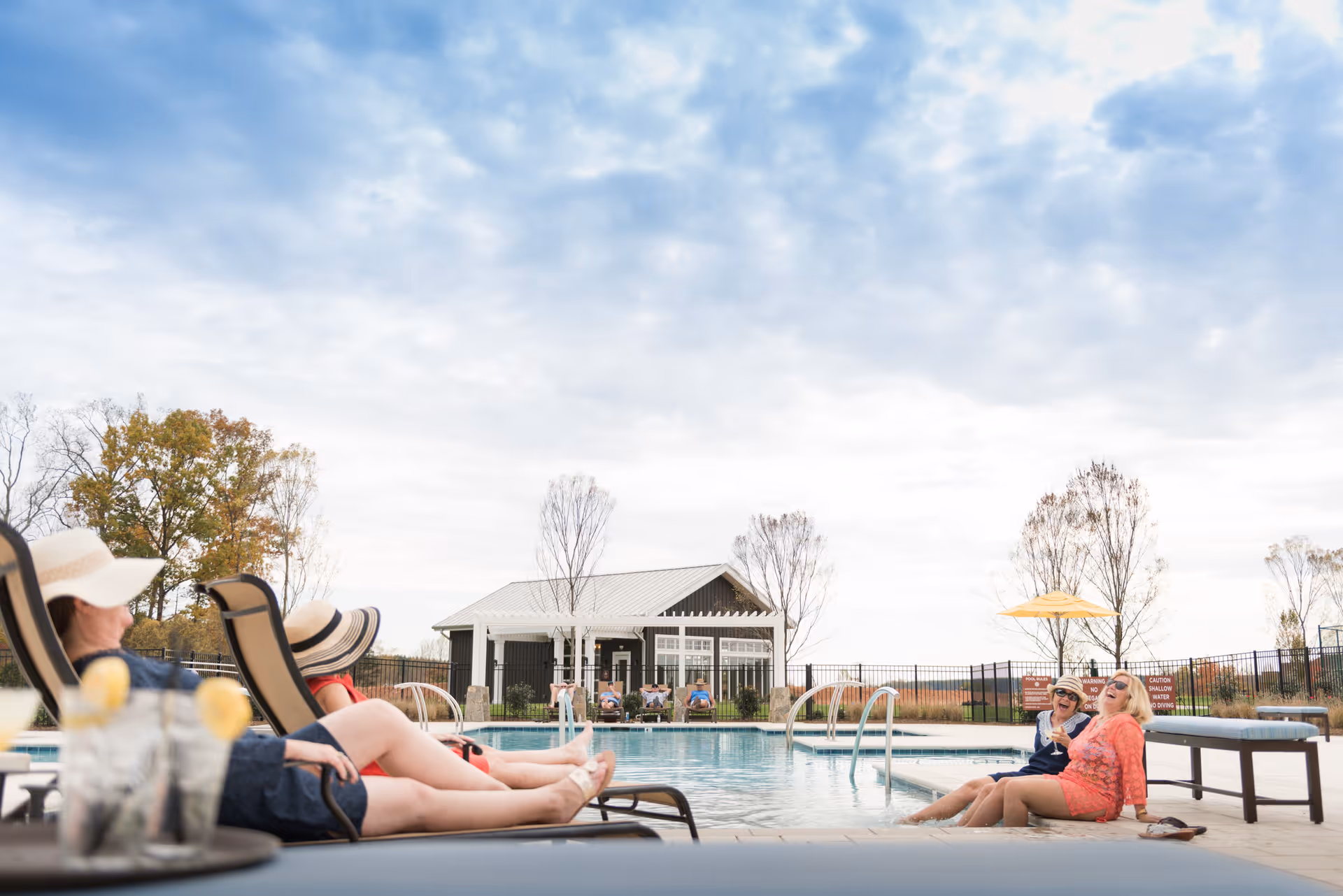 Two women sitting on lounge chairs by an outdoor swimming pool, wearing sun hats and relaxing. Two other women are sitting at the edge of the pool with their feet in the water, laughing and enjoying the sunny day. In the background, there is a small building and some leafless trees under a partly cloudy sky.