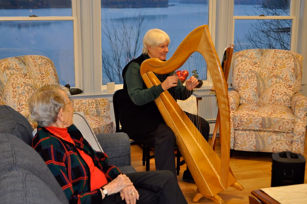 An elderly woman plays a wooden harp in a cozy senior living common room as another resident watches.