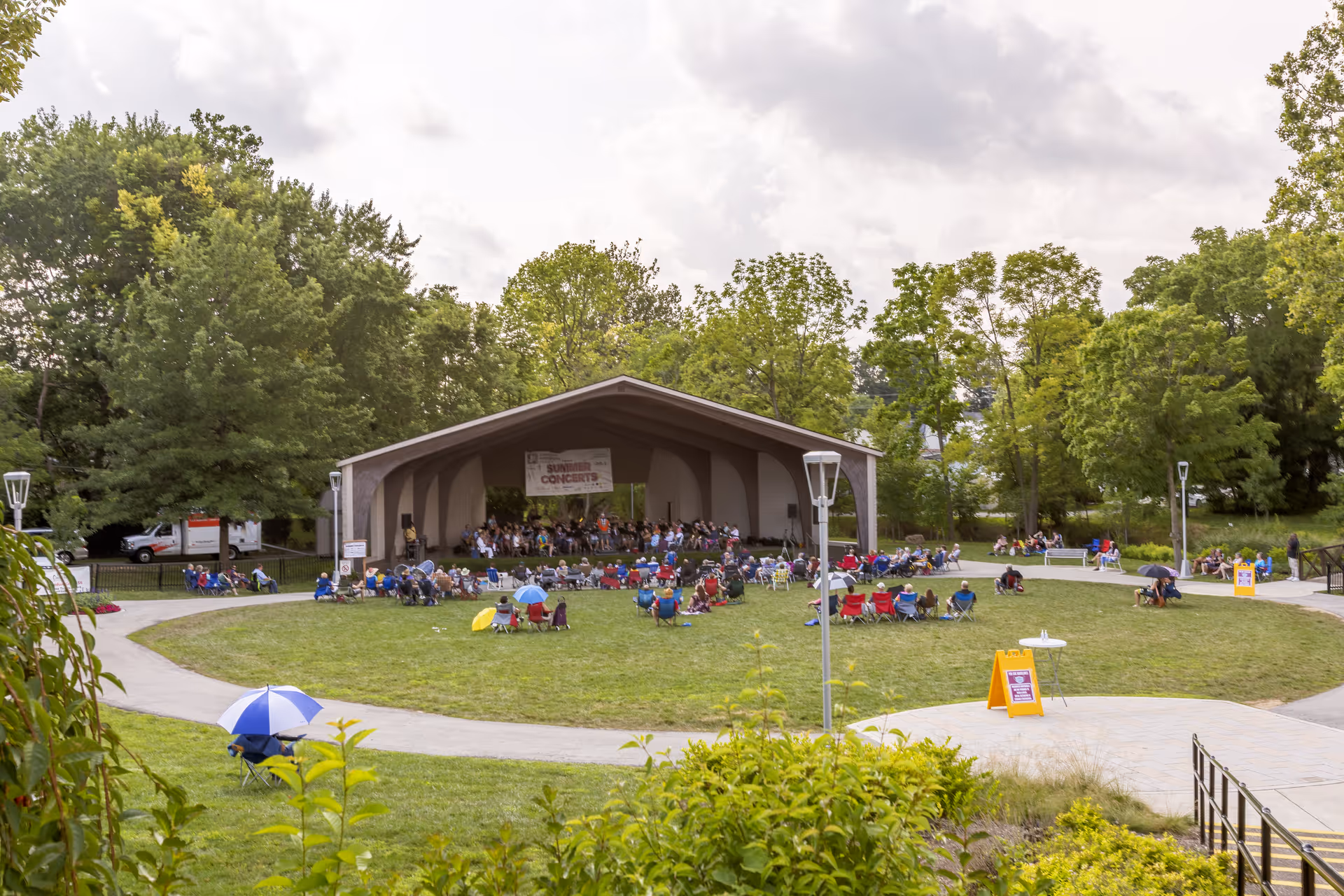 Outdoor amphitheater with a stage where a group of musicians is performing a concert. Audience members are seated on lawn chairs and blankets on the grass, surrounded by trees and greenery under a cloudy sky.