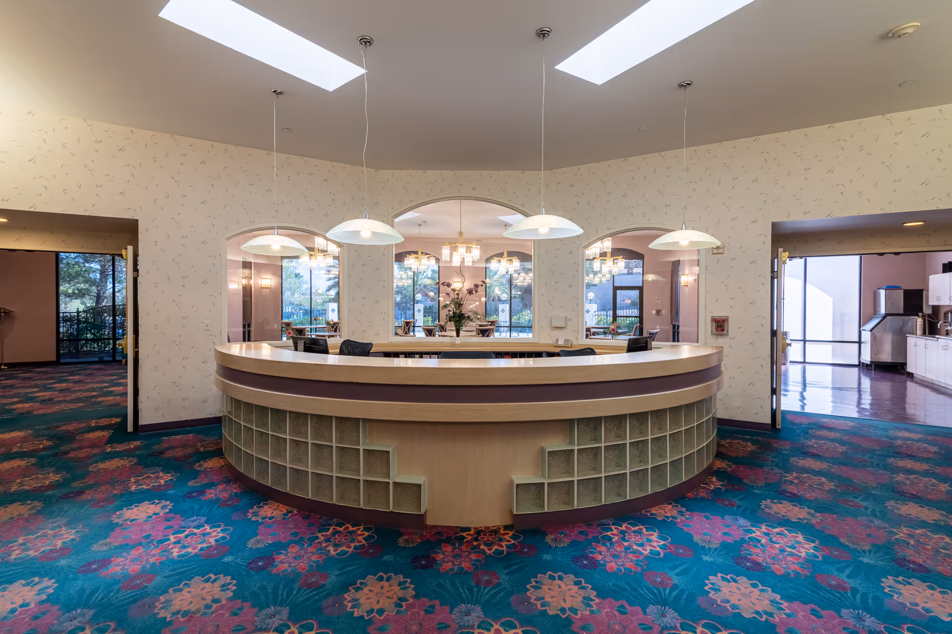 Interior view of a senior living facility reception area with a curved wooden desk featuring glass block accents. The room has a colorful floral carpet, three pendant lights hanging from the ceiling, and arched windows and doorways leading to other rooms with large windows and chandeliers.
